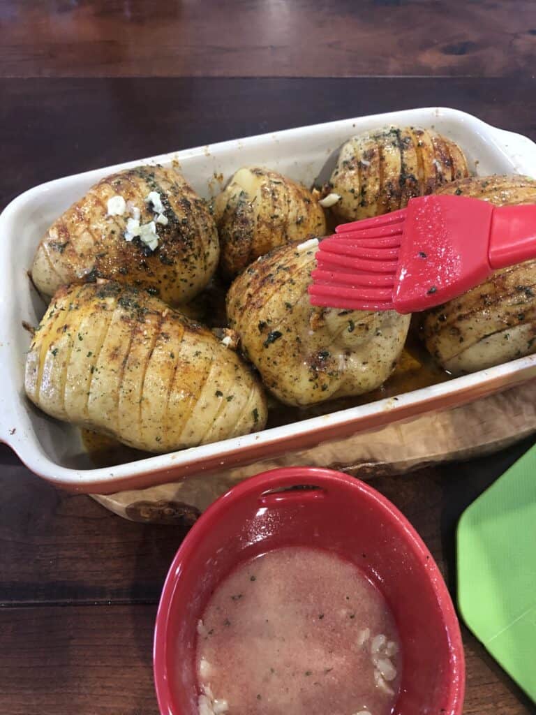 Basting Hasselback Potatoes in a baking pan with red basting brush.