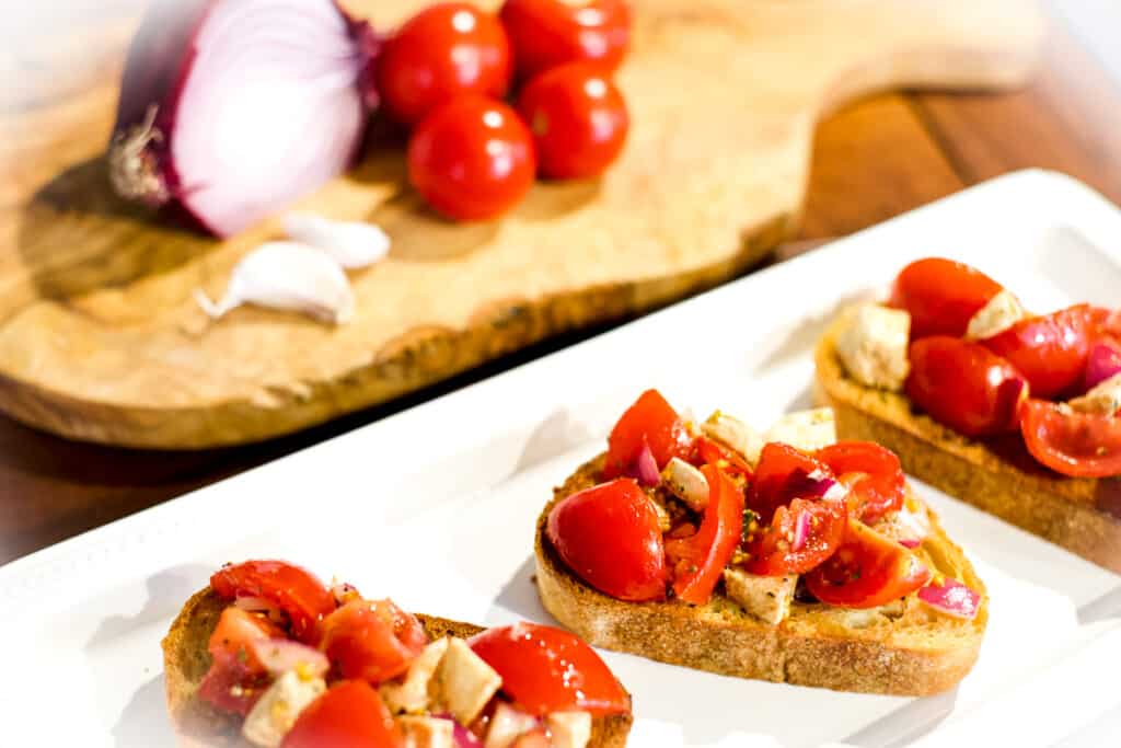 The best caprese bruschetta on a plate with an onion, tomatoes, and garlic off to the side.
